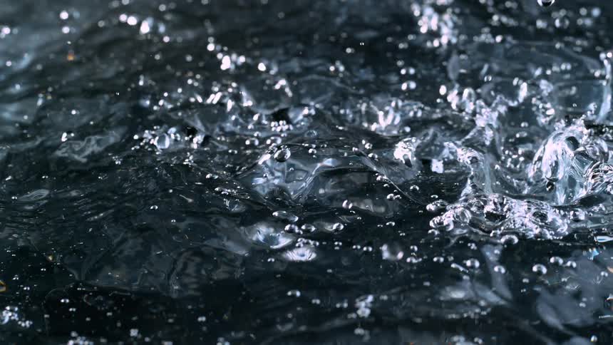 Crystal clear water droplets fall into a dark liquid, creating bubbles and ripples in super slow motion
