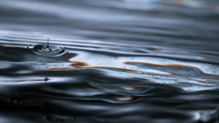 A macro view shows a single drop falling into a dark, viscous, iridescent liquid, creating mesmerizing slow-motion ripples