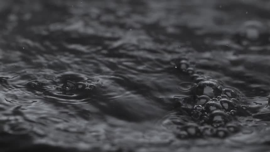 A dramatic black and white slow-motion close-up of raindrops falling onto dark, bubbling water, creating mesmerizing ripples
