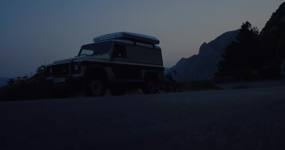 A still shot of an off-road vehicle with a rooftop tent parked on a mountain road at twilight