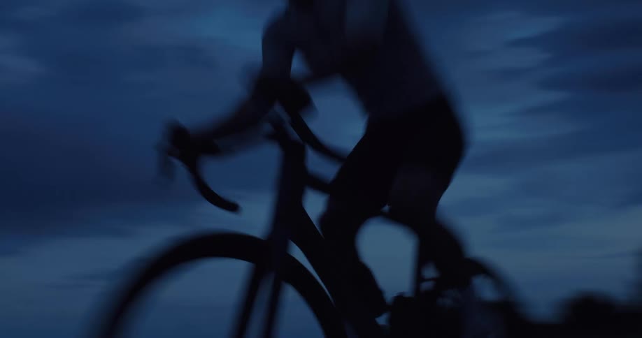 A cyclist's silhouette is captured in a fast-paced, blurred shot as they ride intensely towards the camera against a dark, cloudy sky