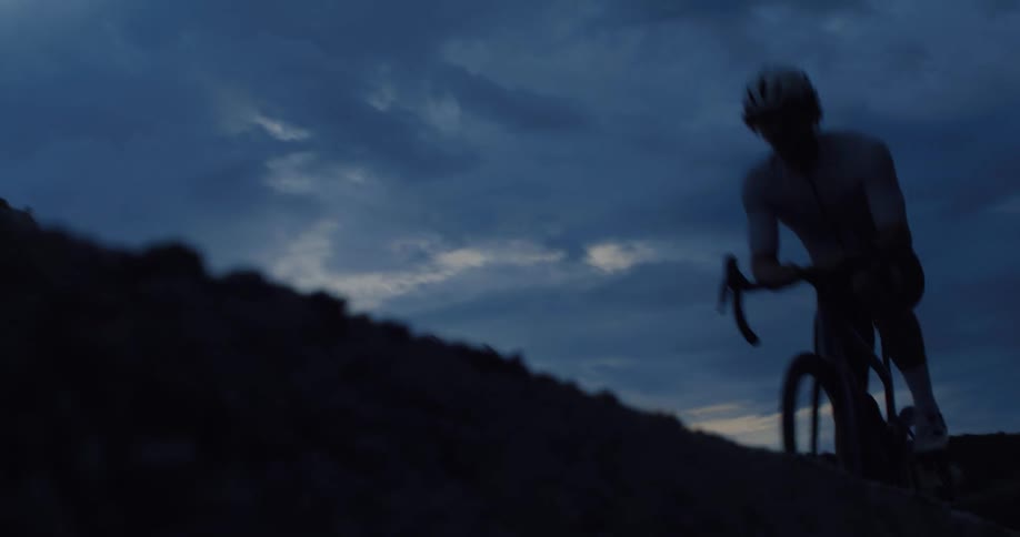 A silhouetted cyclist rides uphill in slow motion against a dramatic, moody sky at dusk