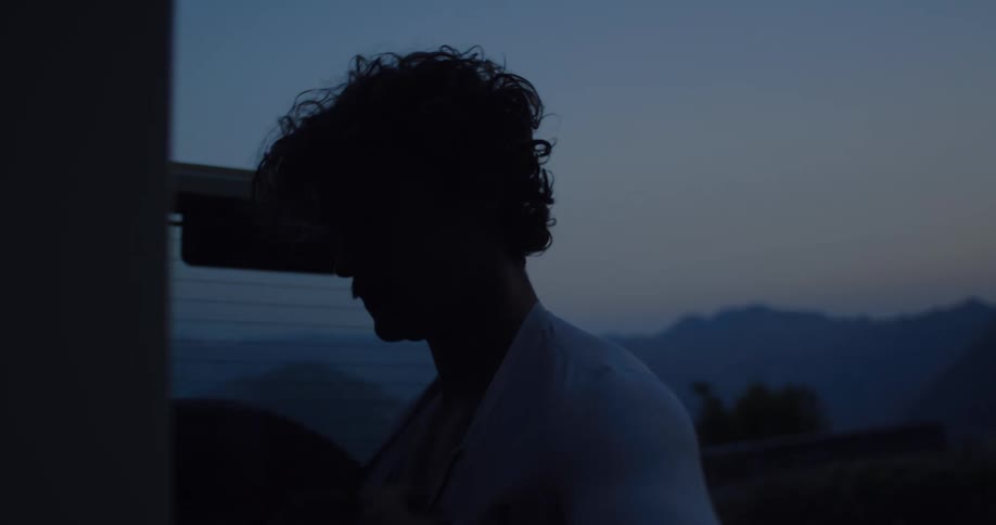 A young man is silhouetted as he slowly gets dressed at dusk, with mountains visible in the fading light