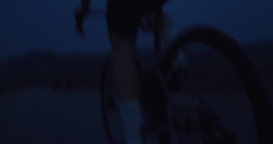 A motion-blurred, low-angle shot follows a cyclist's pedaling legs and spinning wheel on a dark road at night