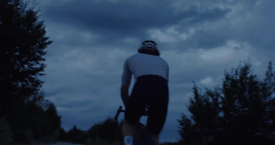 A low-angle shot follows a cyclist wearing a strange ape mask as they pedal down a dark country road at twilight