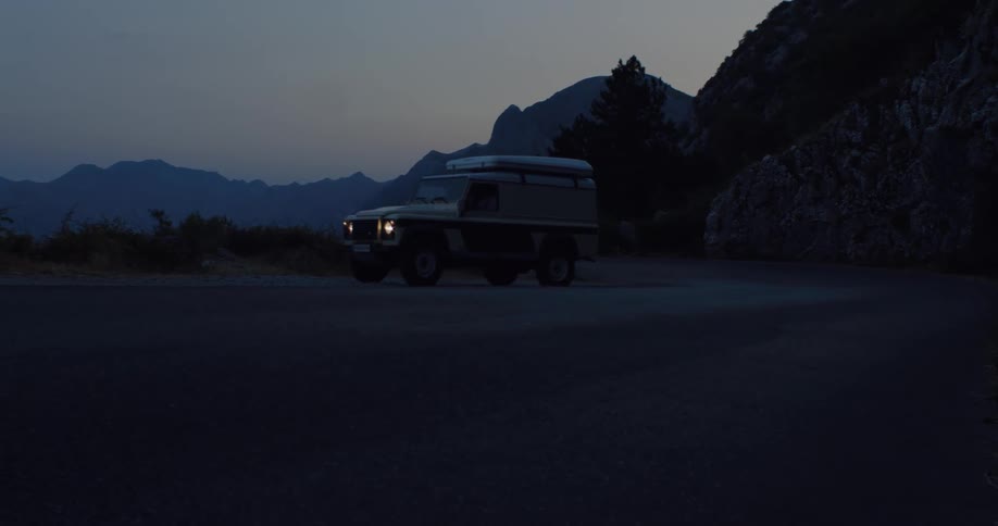 An off-road vehicle with a rooftop tent turns on its headlights while slowly navigating a winding mountain road at dusk
