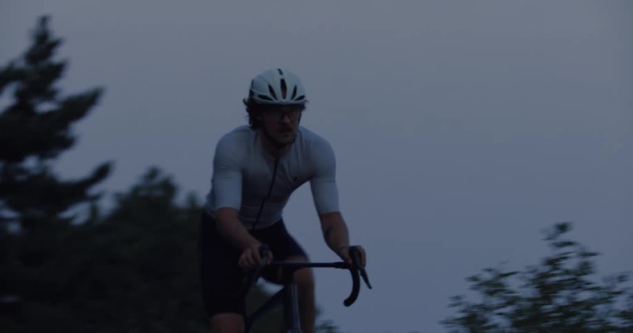 A determined male cyclist in full gear trains on a dark, tree-lined road at dusk