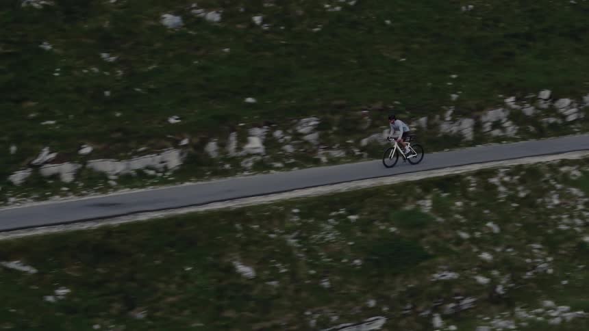 An aerial view follows a cyclist riding along a paved road on a green, rocky mountainside