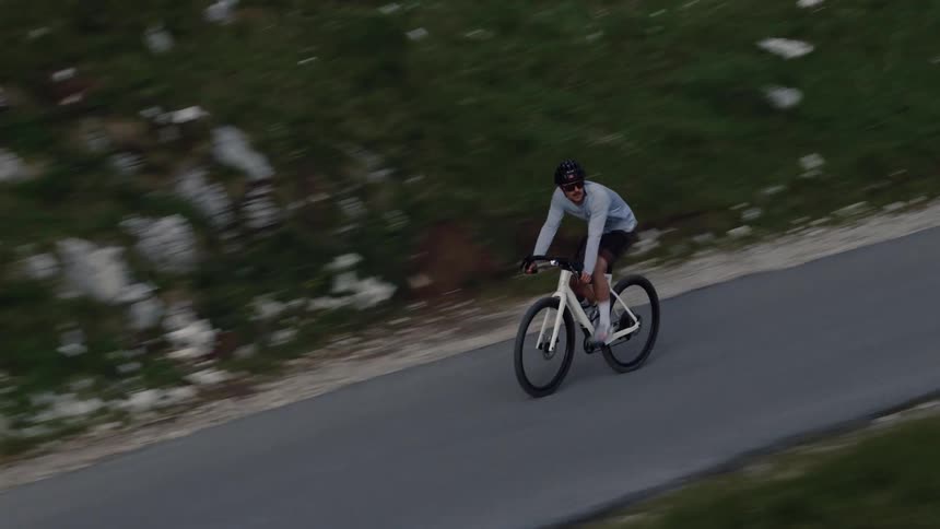 A cyclist in athletic gear pedals up a steep mountain road, captured from a dynamic side-view aerial shot