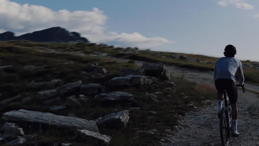 Slow-motion aerial shot follows a man riding his gravel bike along a winding dirt path in a vast, rugged mountain landscape