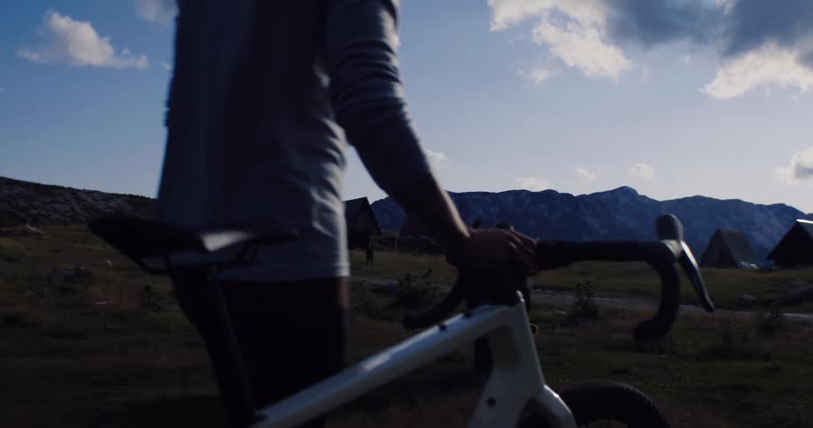 A person in cycling gear walks alongside their bicycle through a scenic mountain pasture with grazing cows and cabins at golden hour