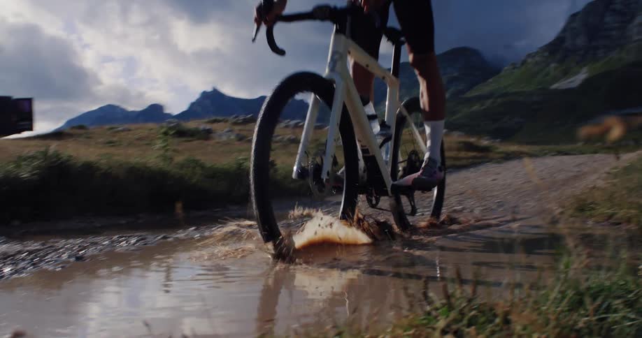 Dramatic slow motion shot of a cyclist riding his gravel bike through a muddy puddle in a stunning mountain landscape