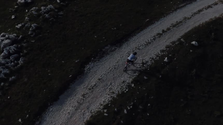 Top-down aerial shot follows a cyclist riding along a winding gravel path in a rugged mountain landscape