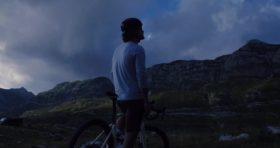 A cyclist pauses to take in the breathtaking mountain views under a dramatic sky before riding off-road into the twilight