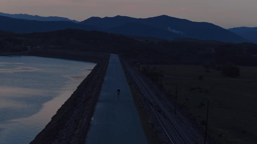 An aerial view shows a lone cyclist riding down a long, straight road on a dam at twilight with mountains in the background