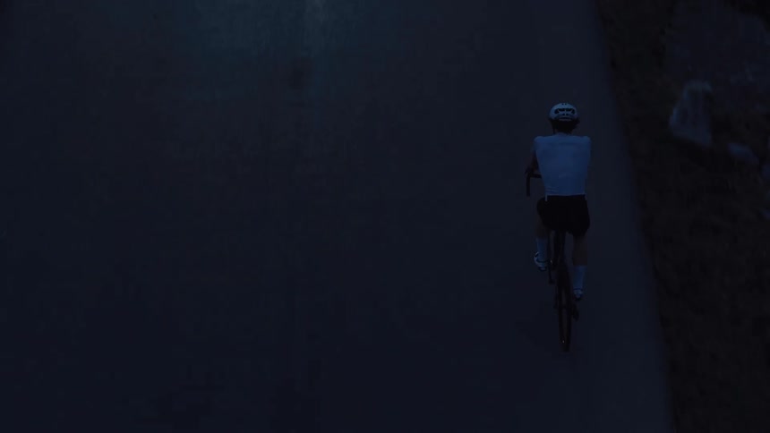 A dramatic top-down aerial shot captures a lone cyclist riding on a dark, paved road at dawn