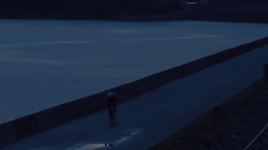 An aerial tracking shot follows a lone cyclist riding on a dark, desolate road next to a lake at dusk