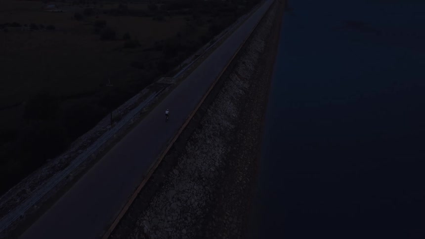 An aerial shot follows a solitary cyclist along a remote dam road at dusk next to a still lake