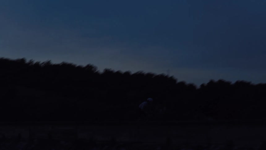 An aerial view follows a silhouetted cyclist riding along a deserted road at twilight