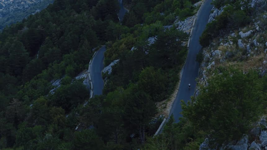 An aerial view shows a lone cyclist tackling a steep, winding mountain road surrounded by a dense forest