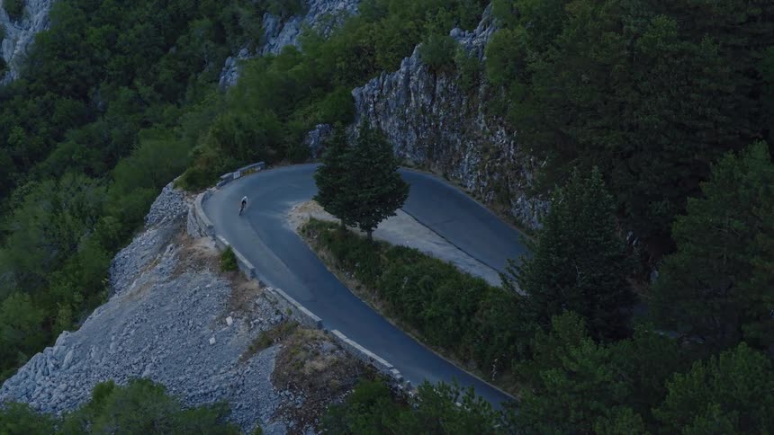 A lone cyclist tackles a steep hairpin turn on a winding road through a dense mountain forest