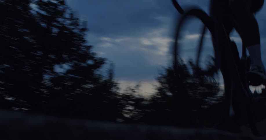 A cyclist rides quickly past the camera on a country road at dusk, silhouetted against a dark, cloudy sky