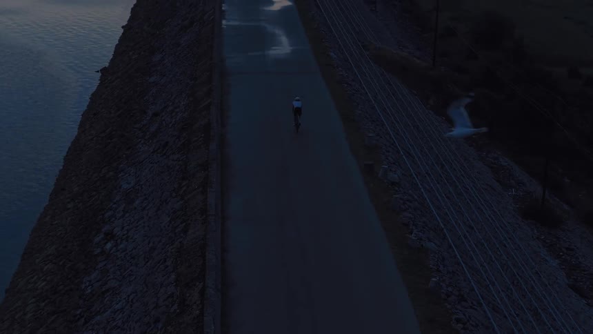 An atmospheric aerial view shows a lone cyclist riding along a lakeside dam at dusk as a bird flies through the frame