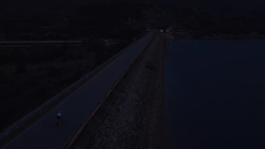 An epic aerial shot follows a lone cyclist riding on a road over a large dam at dusk