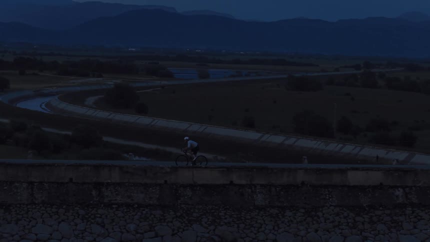 A lone cyclist rides along a high dam road at twilight, with a winding river and mountains in the background