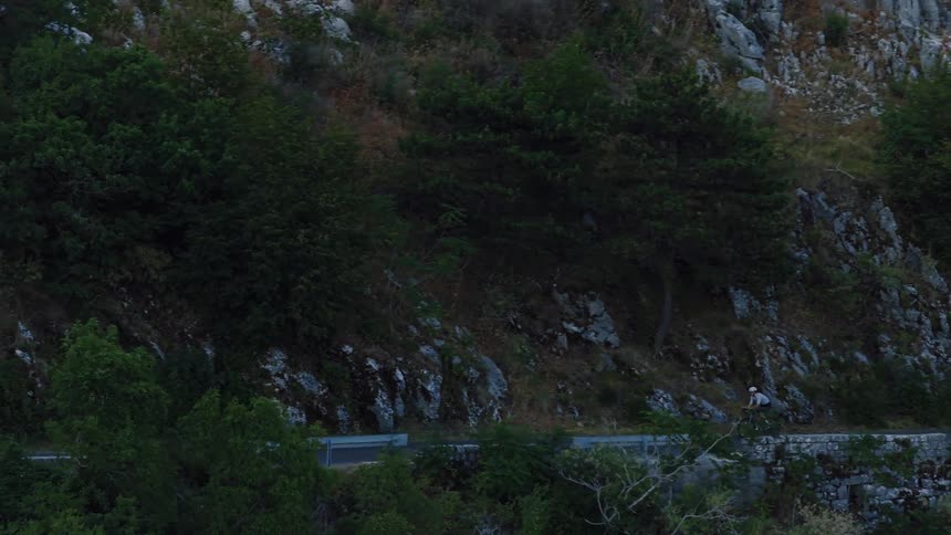 An aerial shot follows a determined cyclist riding along a steep mountain road at dusk
