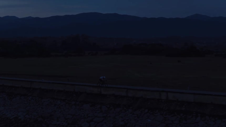An aerial view follows a lone cyclist riding along a dam road next to a large lake in the mountains at dusk