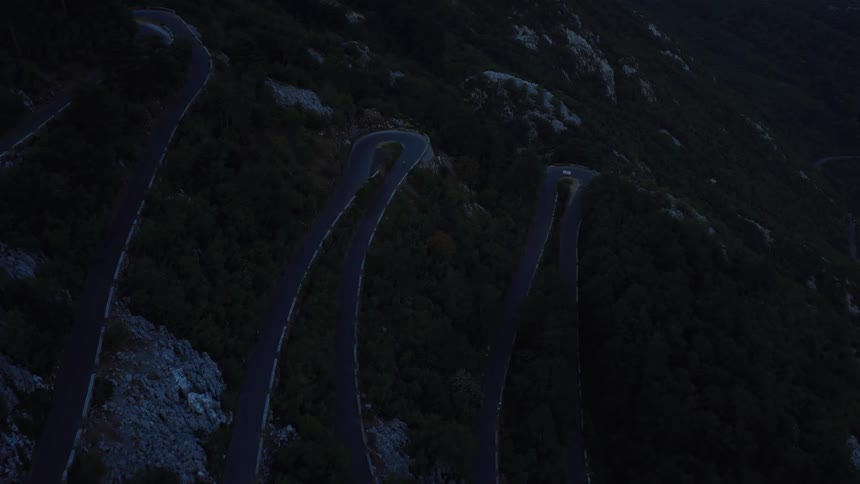 A lone vehicle carefully navigates the sharp hairpin turns of a winding mountain road at twilight