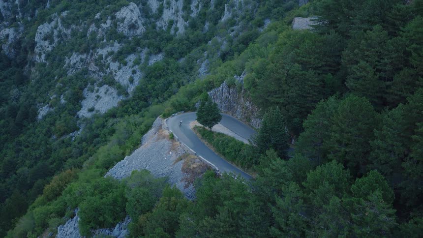Epic aerial shot shows a single cyclist making their way around a steep hairpin turn on a mountainside road through a lush forest