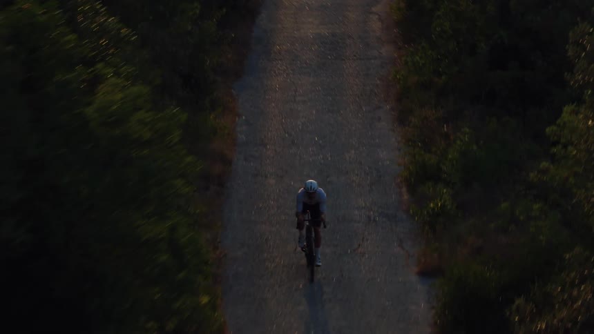 Aerial top-down view of a cyclist riding uphill on a secluded forest road during a golden hour sunset