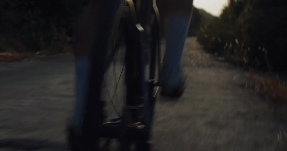 Dynamic low-angle shot of a cyclist's legs and rear wheel spinning on an asphalt road at sunset