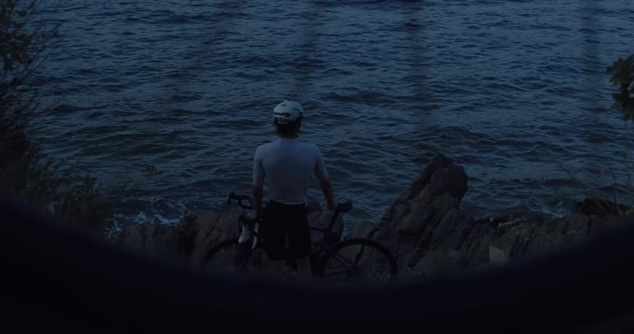 A cyclist with his bicycle stands on a rocky coastline at dusk, looking out over the dark blue water