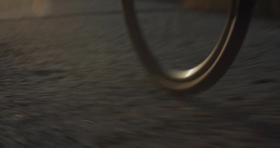 A low-angle POV shot captures the wheels of a bicycle spinning as a person pedals down an asphalt road during a beautiful sunset