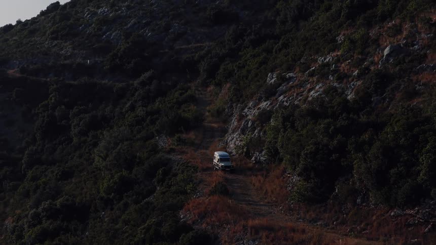 Aerial view slowly rises above an off-road camper van parked on a remote dirt trail in the rugged, forested mountains at sunset