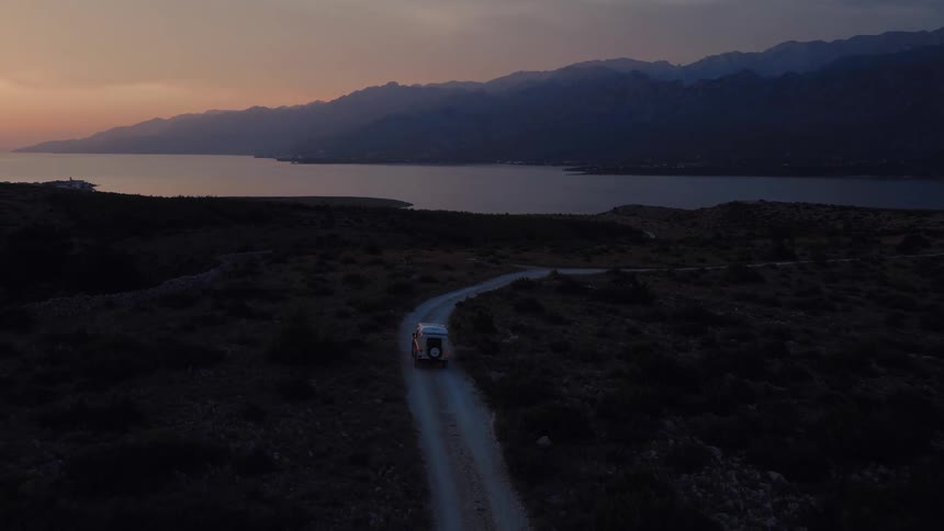 An aerial view shows a camper van driving on a scenic dirt road along the coast with mountains in the background at dusk