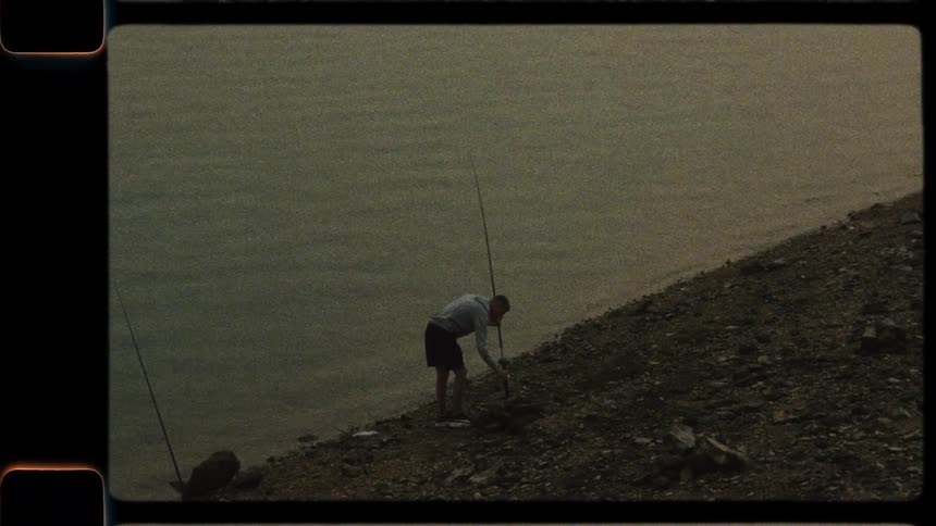 A lone fisherman sets up his rod on the rocky bank of a serene lake in this vintage-style footage