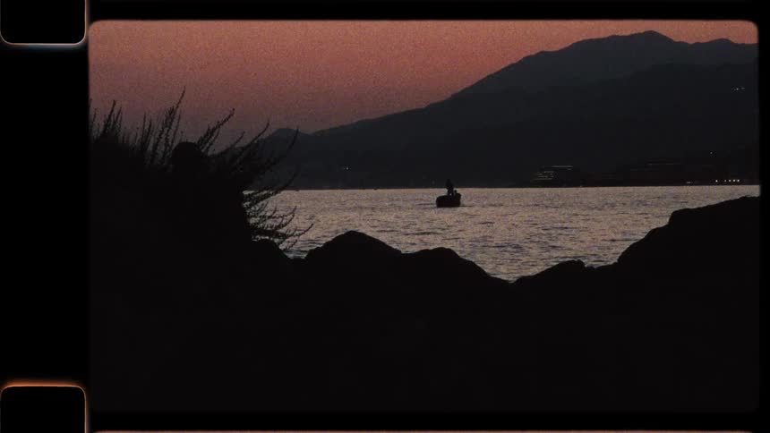 A vintage Super 8 film captures a tranquil scene of a lone boat silhouetted on the water at sunset, with mountains in the background
