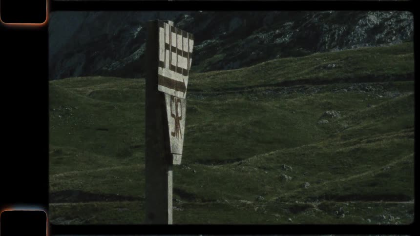 A vintage film shot of a rustic trail marker standing alone in a vast, green mountain landscape