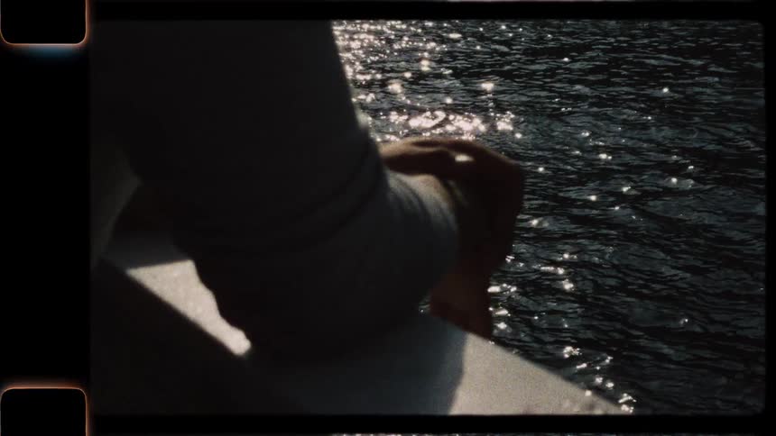 A close-up vintage 8mm film shot shows a person's hands silhouetted against the sparkling sunlit water