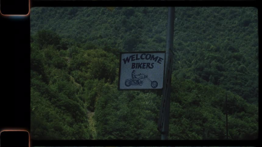 A vintage film shot of a 'Welcome Bikers' sign against a dense forest, panning up to Montenegrin flags