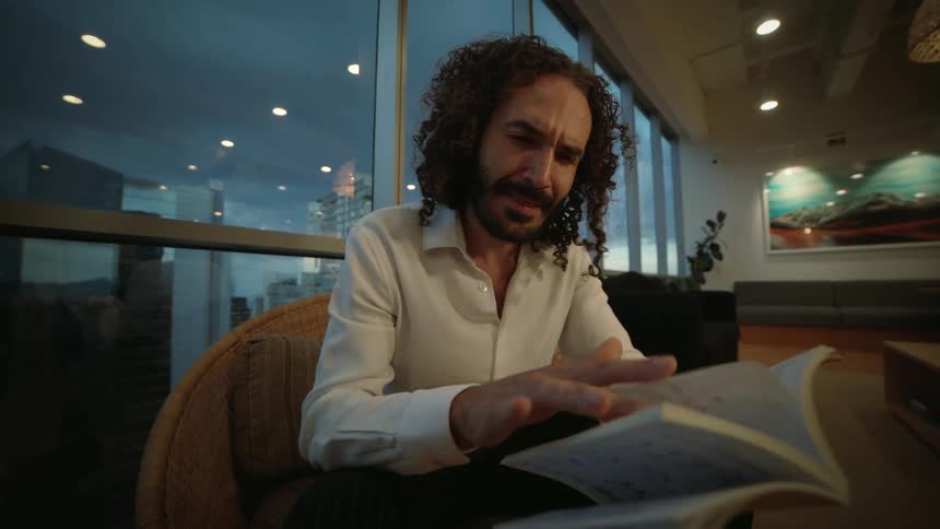 A handsome man with curly hair reads a book intently while sitting in an armchair in a modern high-rise lounge at dusk