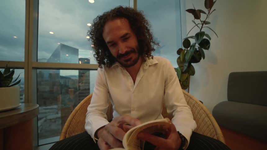 A man with curly hair enjoys reading a book while sitting in a stylish chair in a modern high-rise apartment with a city view