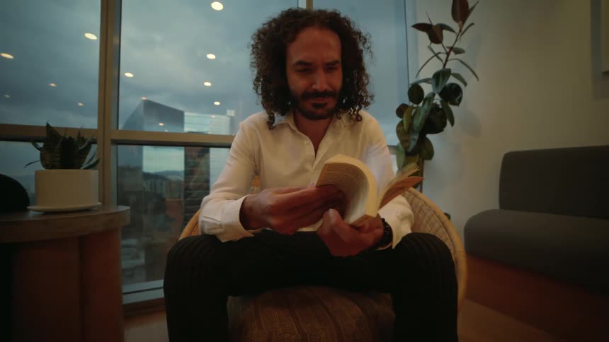 A handsome man with curly hair sits in a modern chair and flips through a book in slow motion in a high-rise apartment