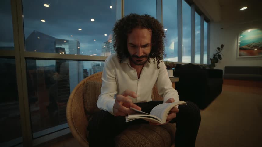 An intense man with long curly hair reads a book in a modern room with a large window overlooking the city at dusk