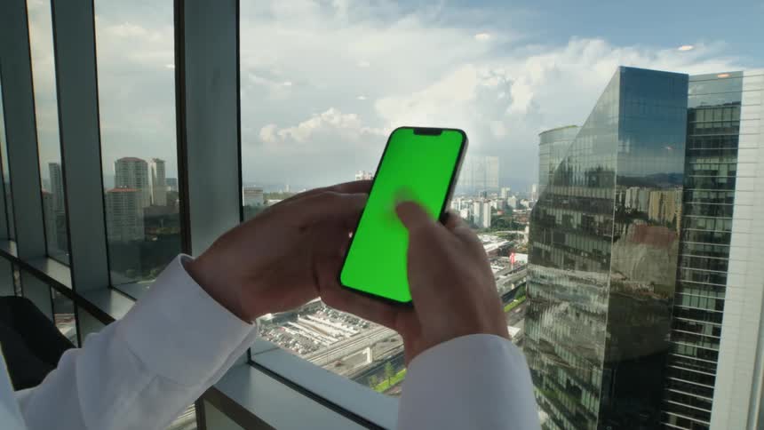 A businessman in a white shirt scrolls on a green screen smartphone in front of a window with a city view