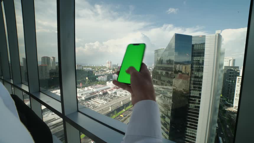 A businessman in a modern high-rise office swipes on a green-screen smartphone with a sprawling cityscape view